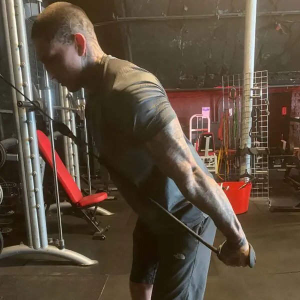 A tattooed individual exercises in a gym, using TriGrips Long Handles on a cable machine for tricep pushdowns. The gym features diverse equipment like a red bench and weights, all set against dark floors and ceilings.