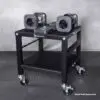 The Ironmaster 75 LB Quick-Lock Adjustable Dumbbell Set, without the stand, is organized on a black wheeled rack featuring two levels: dumbbells on top and weight plates below. The floor has a textured rubber mat pattern.
