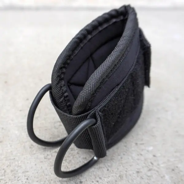 The Spud Ankle Cuff Pair features a close-up of a black fabric strap with two D-rings on a concrete surface, showcasing visible stitching and a quilted pattern on one side, complemented by Velcro material on the other.
