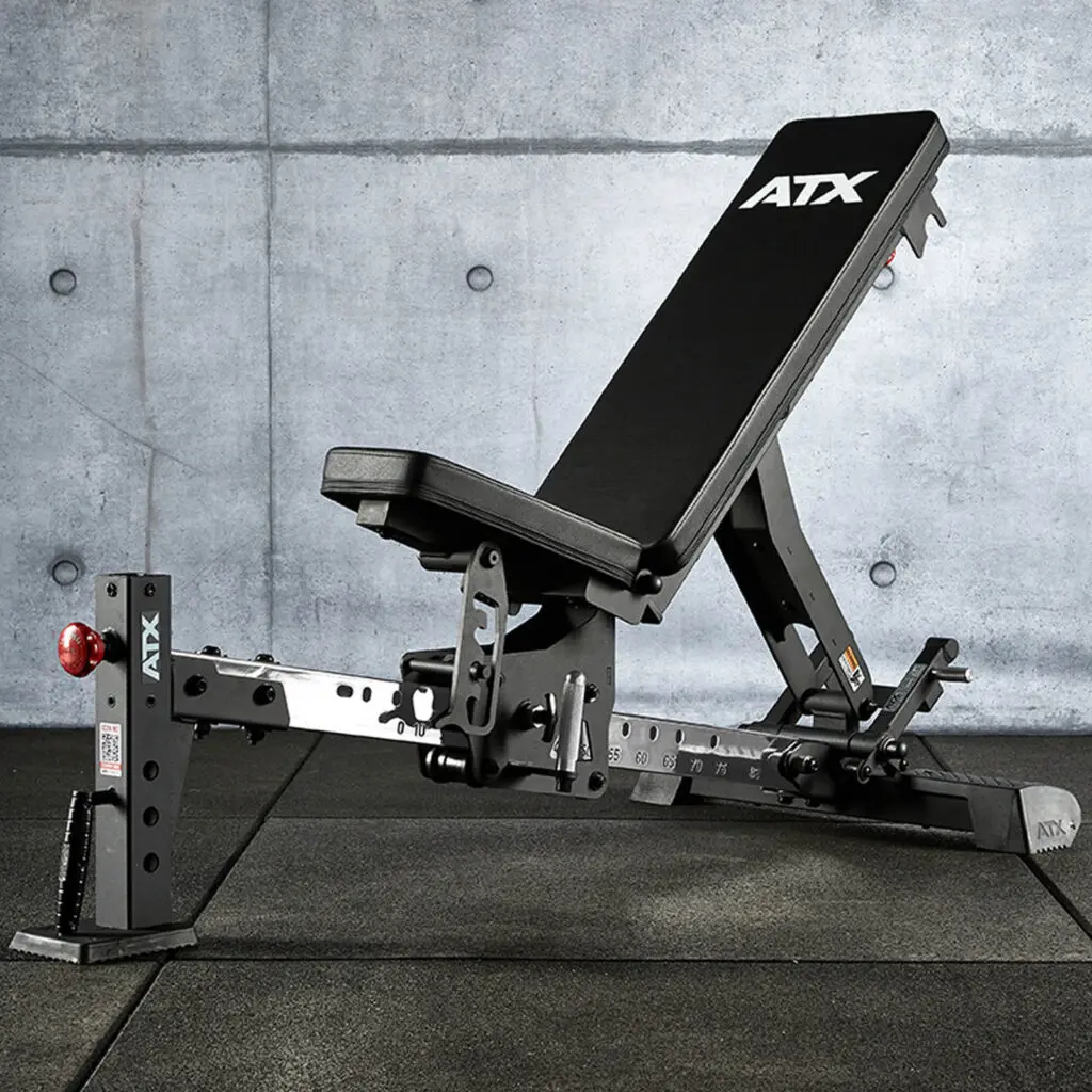 A black FID bench with multiple incline settings is positioned on a dark gym floor. The background features a textured concrete wall, enhancing the industrial aesthetic. The bench is labeled ATX in crisp white lettering.
