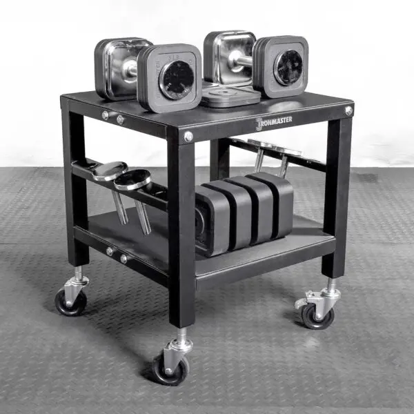 The Ironmaster QLDB Stand PRO showcases a modern black design with square-shaped adjustable dumbbells on wheels. It features two shelves for extra weights and accessories, all sitting on a durable rubber mat.