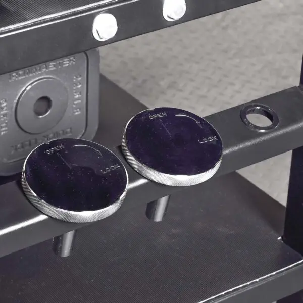 Close-up of two round, dark knobs labeled OPEN and LOCK on the Ironmaster QLDB Stand PRO, attached to a black metal frame above a textured gray floor.