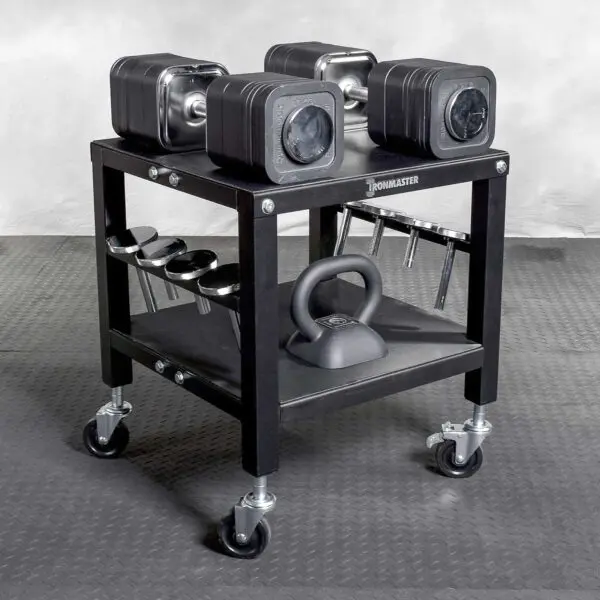 The Ironmaster QLDB Stand PRO, a black wheeled rack, neatly holds adjustable dumbbells and a kettlebell on a textured gray floor. It has extra weights and includes a compartment for weight plate storage.