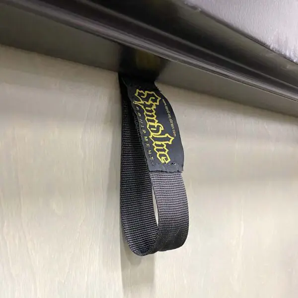 Close-up of a black Spud Inc Door Jammer strap featuring Sound Line in yellow lettering, attached to a horizontal metallic surface, set against a textured wall.