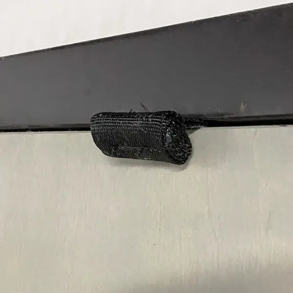 Close-up of a black Velcro strap from the Spud Inc Door Jammer attached to a two-toned metal surface, secured with a metal bracket. The fabric at the edges appears slightly frayed.