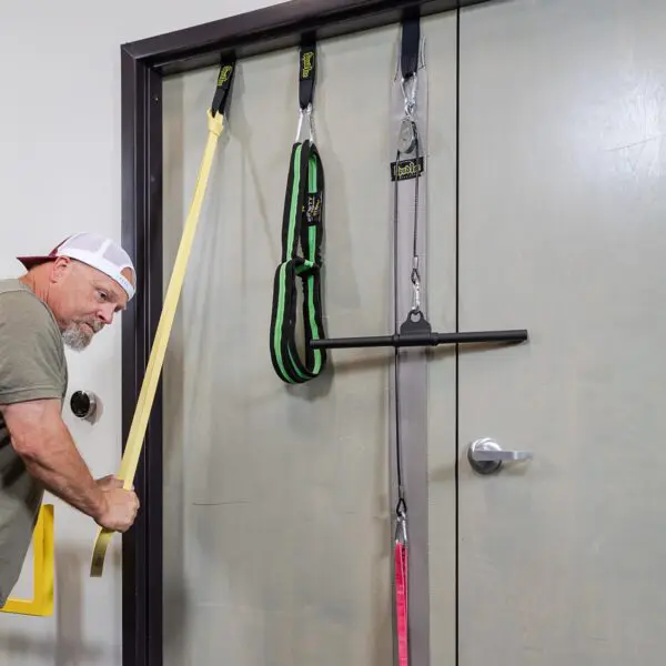 A man in a cap and khaki shirt uses a yellow resistance band with the Spud Inc Door Jammer, which also includes green and gray bands for diverse workouts secured on a closed door.