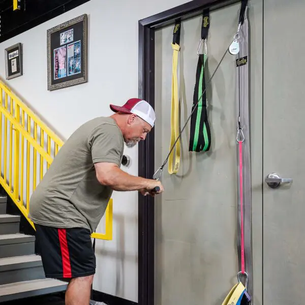 A man in a backward baseball cap and athletic wear exercises with the Spud Inc Door Jammer Pulley System attached to a door. Hes near a staircase with yellow railings, and framed pictures adorn the wall.