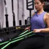 A woman in a purple tank top sits on the gym floor, using a green Spud Inc Infinity Loop for resistance exercises. She is focused and performing a seated pull exercise with gym equipment in the background.