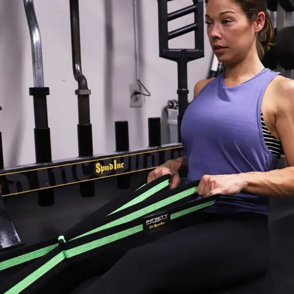 A woman in a purple tank top sits on the gym floor, using a green Spud Inc Infinity Loop for resistance exercises. She is focused and performing a seated pull exercise with gym equipment in the background.