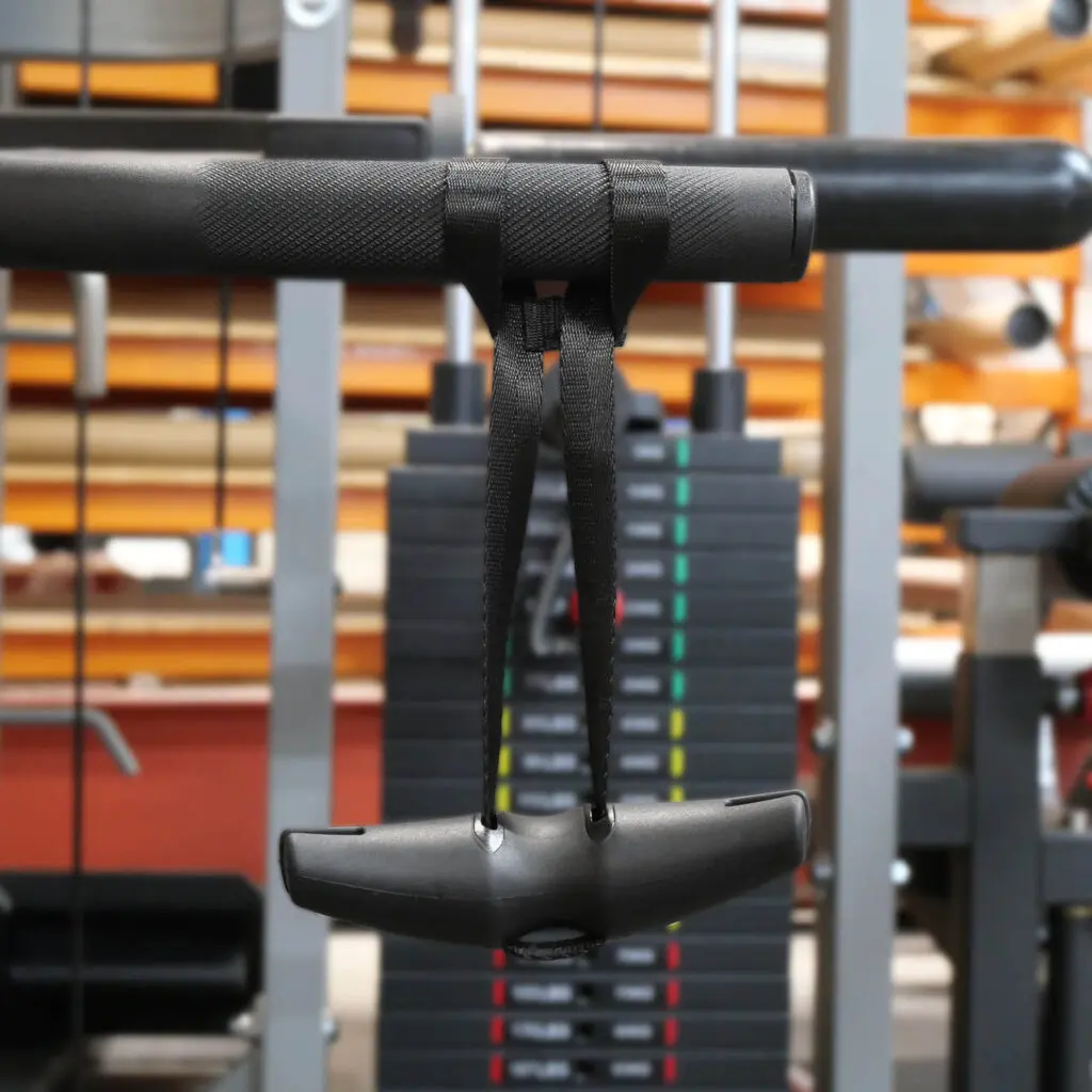 Close-up of a D Angle Grip V2, showcasing its black grip with adjustable straps. The background includes weight stacks and gym equipment, suggesting a fitness environment.