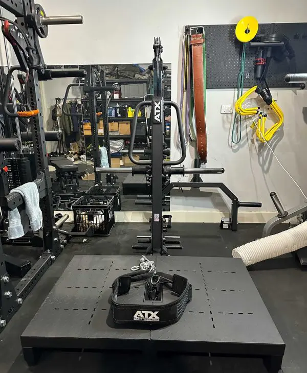 A home gym setup with various exercise equipment including a squat rack, barbells, and belts. A workout belt is placed on a platform in the foreground, and resistance bands hang on the wall. A mirror reflects the equipment.