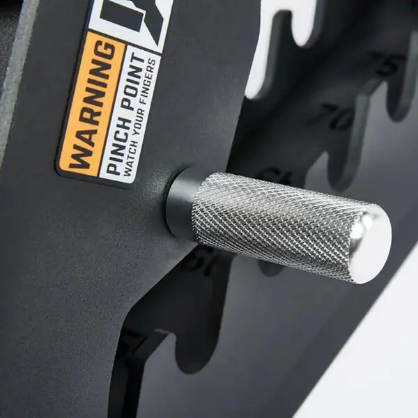 Close-up of a metal handle with textured grip on a black weightlifting machine near the ATX® Commercial FID Bench ATX-MBX-660. It has a warning label: Pinch Point, Watch Your Fingers. Part of the numbered weight stack is visible.