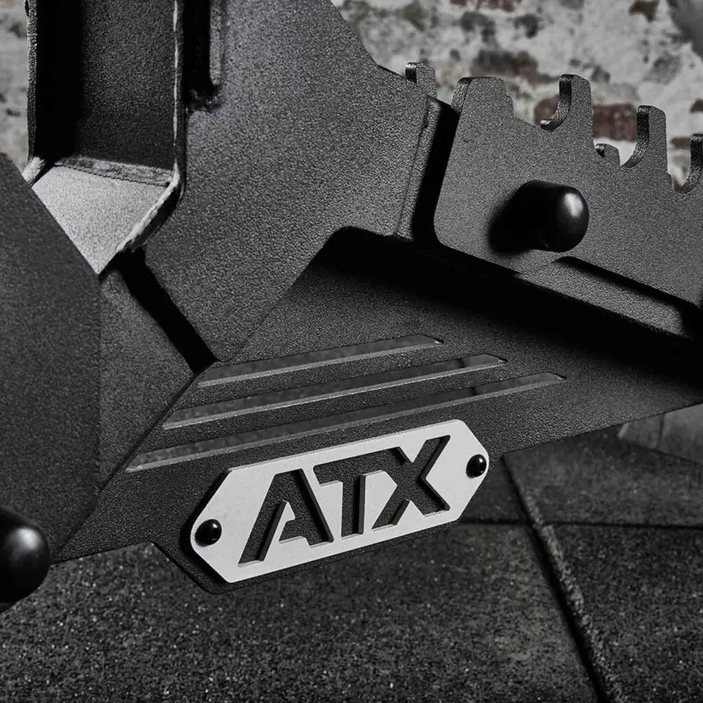 Close-up of a black metal gym equipment piece, featuring a silver plate displaying the letters ATX. An adjustable weight bench forms a textured surface in the background.
