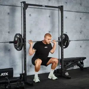 A person, in a black ATX t-shirt and shorts with white socks and light shoes, performs a squat with a barbell on their shoulders inside a gym featuring an ATX® Smith Machine 500 for low ceilings (198 cm), with a weight bench and other equipment visible in the background.