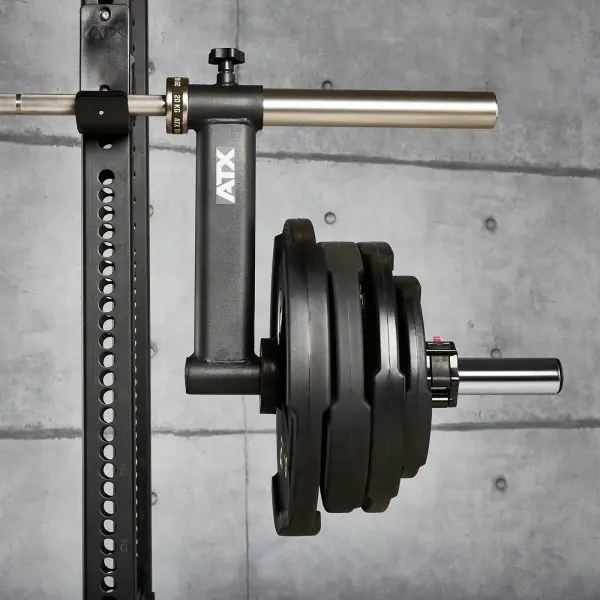 A close-up of a gym equipment piece with several weight plates loaded onto a horizontal bar. The metal frame is attached to a squat rack against a concrete wall. The equipment is labeled ATX.