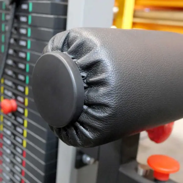 Close-up of a gym machines padded, cylindrical armrest, featuring a black Foam Roller Cover 65mm. A stack of adjustable weights is visible in the background, with numbered markings and colored lines next to red adjustment knobs.