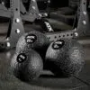 Three ATX® Slam Balls in black texture (4 kg, 12 kg, 20 kg) rest on the gym floor with a metal exercise rack seen behind.