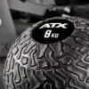 Close-up of a black ATX® Slam Ball labeled 8 KG with a textured surface, set against a blurred gym background.