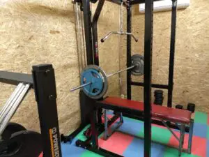 A home gym with a squat rack, a barbell loaded with weights on a bench press, a red bench, rubber floor tiles, stacked weight plates, and an air conditioner mounted on a wood-paneled wall.