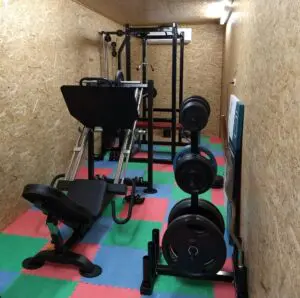 A compact home gym with a leg press machine, weight plates on a rack, a multigym station, and colorful interlocking floor mats covering the floor. The walls are paneled with light wood.