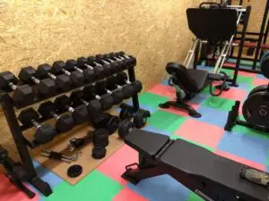 A gym with a rack of hexagonal dumbbells, a bench, various free weights on the floor, and a leg press machine, all set on colorful interlocking floor mats against a wooden wall.