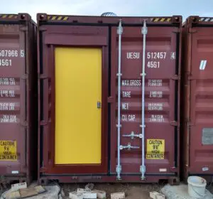 A red shipping container with a bright yellow door installed in the center. Warning labels read CAUTION 96 HIGH CONTAINER. The container is surrounded by wooden blocks and sits on a rocky surface.
