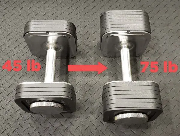 Two Ironmaster adjustable dumbbells rest on a black textured floor, with the left labeled 45 lb and the right labeled 75 lb. A red arrow points from the left dumbbell to the right, suggesting progression in your workout.