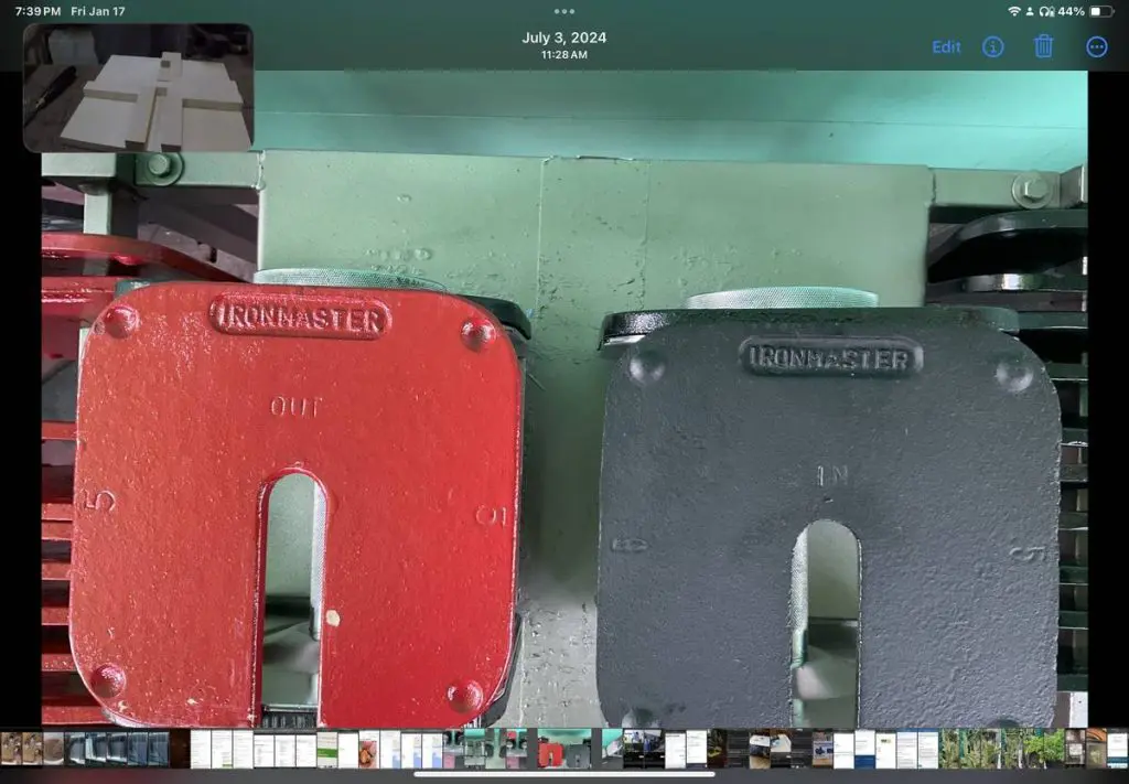 Close-up of two weight plates labeled IRONMASTER on Eisenlink Dumbbells. The red plate is marked OUT, and the black plate is marked IN. Both are attached to metal equipment, viewed from above.