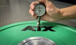 A hand holds a durometer gauge measuring the hardness of a green ATX weight plate resting on a metallic surface.