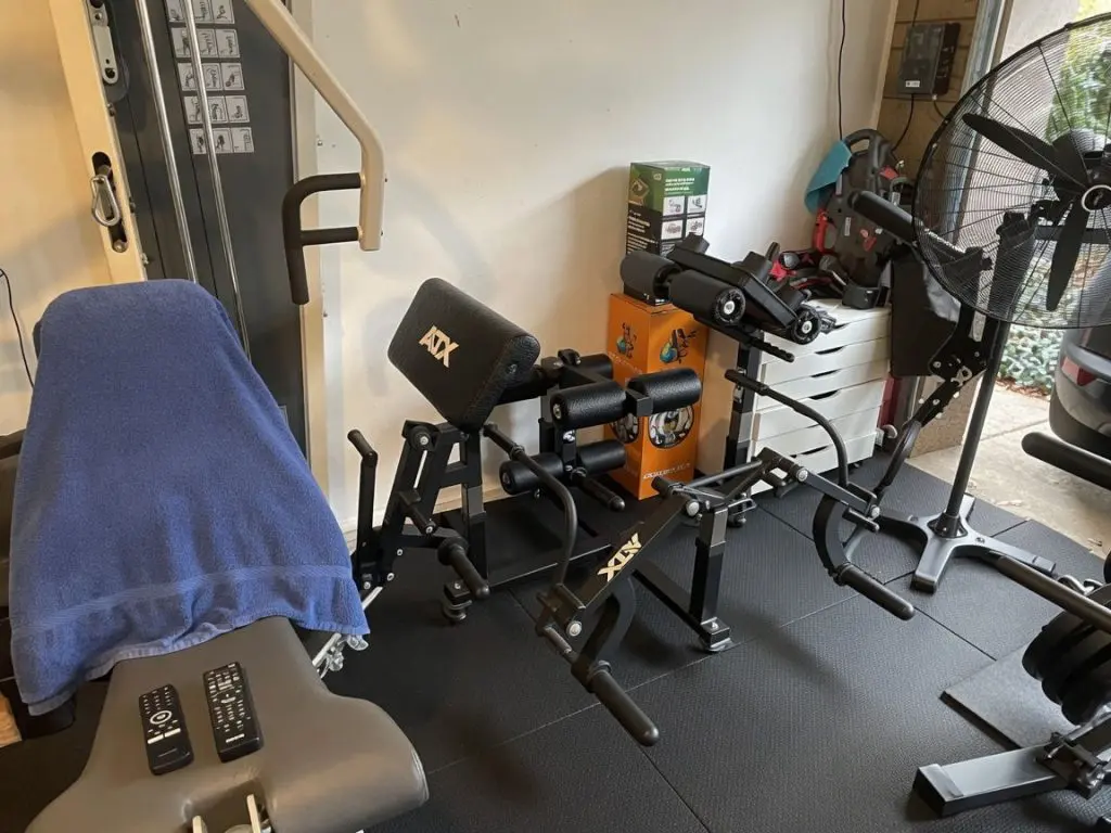 A Dans Single Car Garage Gym setup with various exercise machines, a padded bench covered by a blue towel, remote controls on the bench, and a large standing fan nearby. Workout mats cover the floor and windows show greenery outside.