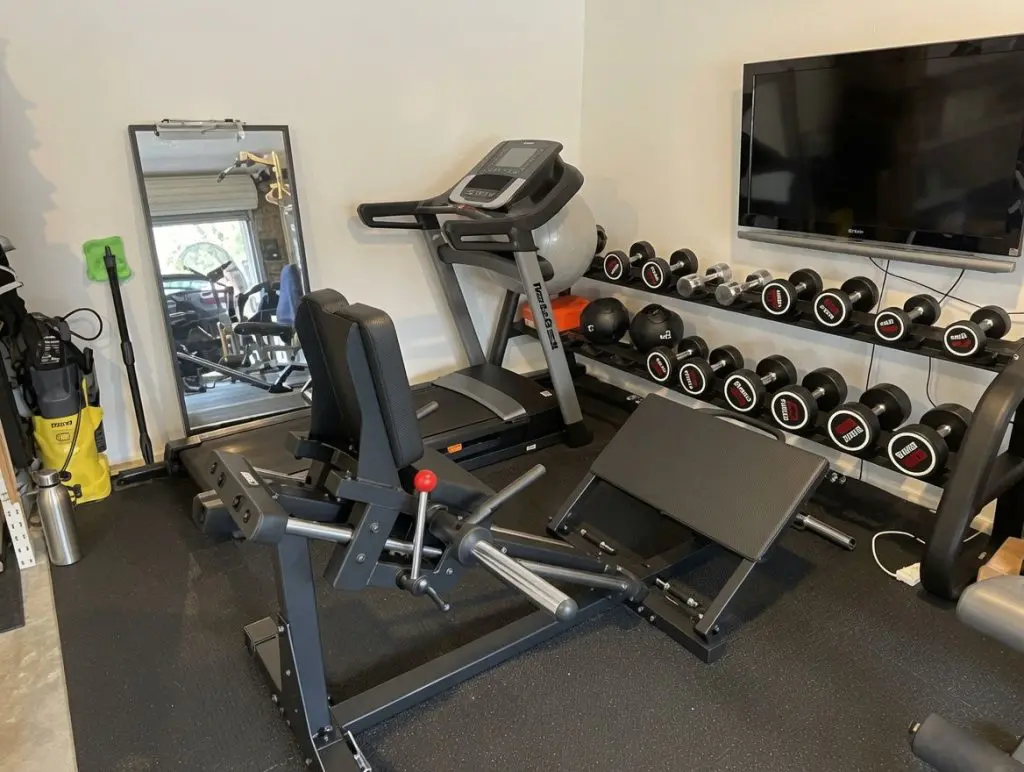 A home gym with a treadmill, leg press machine, wall-mounted TV, mirror, and shelves holding various dumbbells. Golf clubs and cleaning tools are seen in the corner near a window.