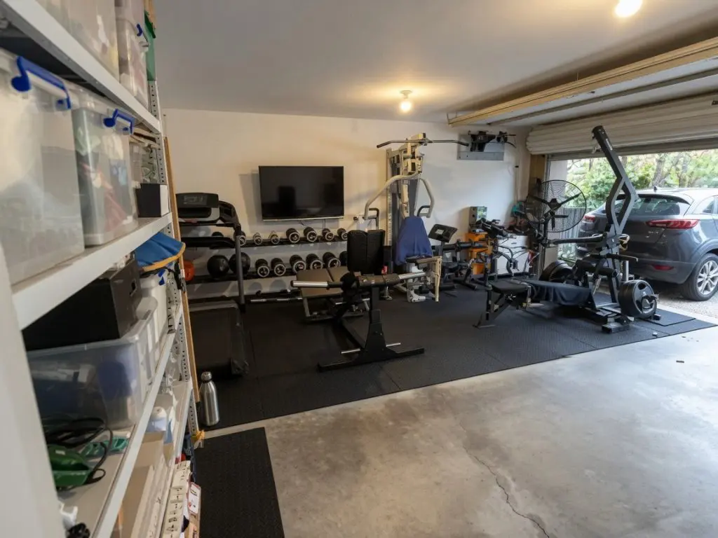 A home gym setup in a garage with various exercise equipment, including weights, benches, machines, a treadmill, and dumbbells. Shelves with storage bins line the left wall, and a parked car is visible on the right.