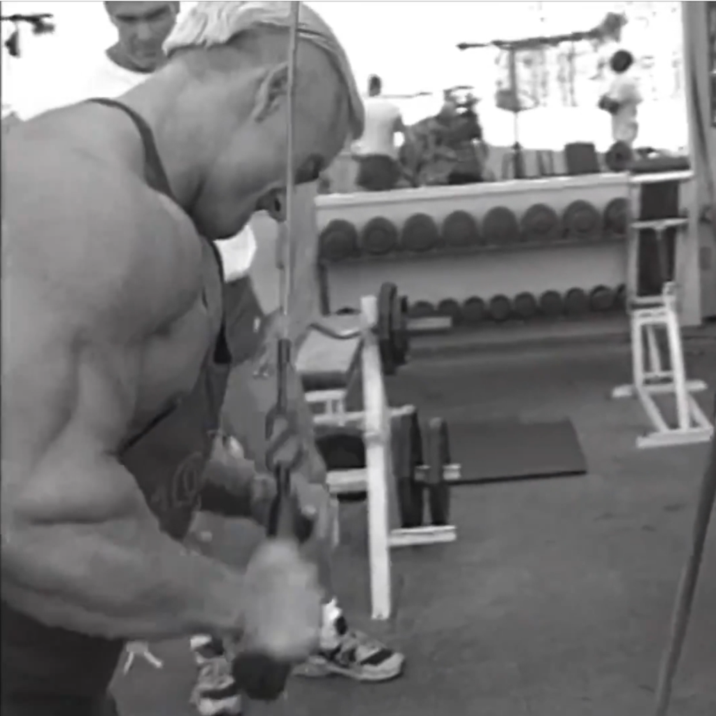 A muscular person does a tricep cable pushdown in a gym, facing sideways. Gym equipment, free weights, and other people are visible in the background. The image is in black and white.