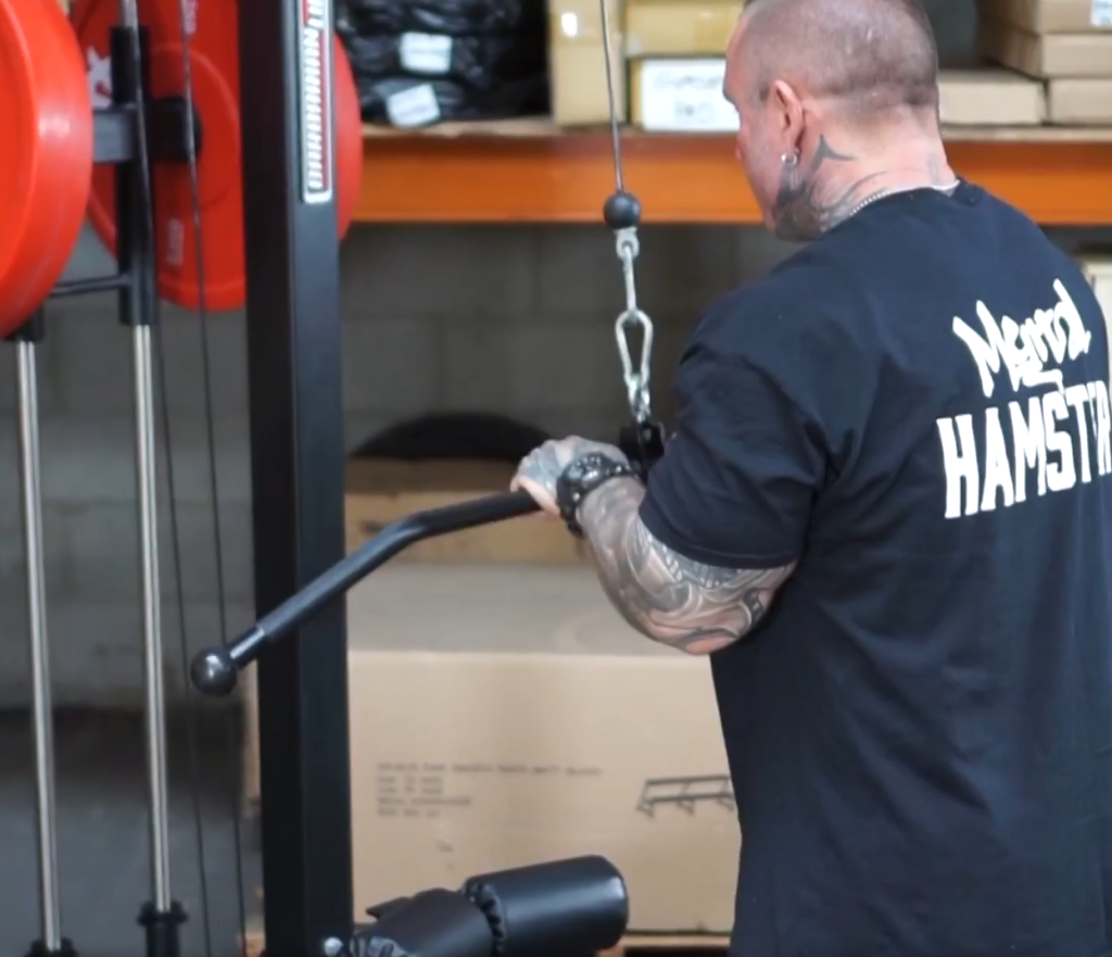 A tattooed man wearing a black t-shirt performs a lat pulldown exercise on a cable machine.