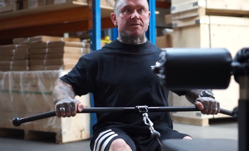 A tattooed man in a black t-shirt and shorts performs a seated cable row exercise in a warehouse.
