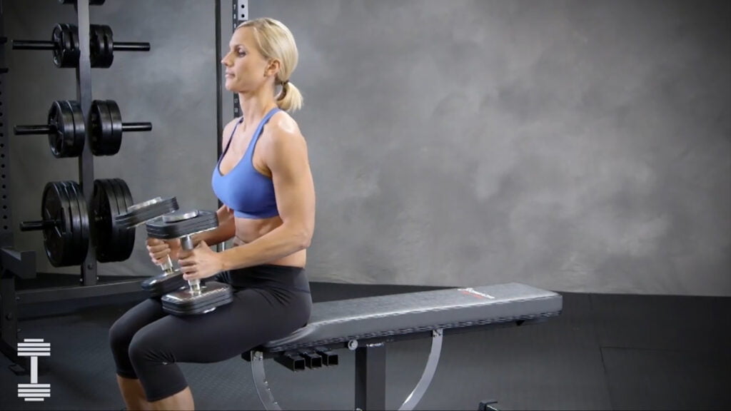 A person in workout attire is sitting on a bench in a gym, holding dumbbells in a hammer curl position. There are weight racks in the background. The scene is set against a plain, gray wall.