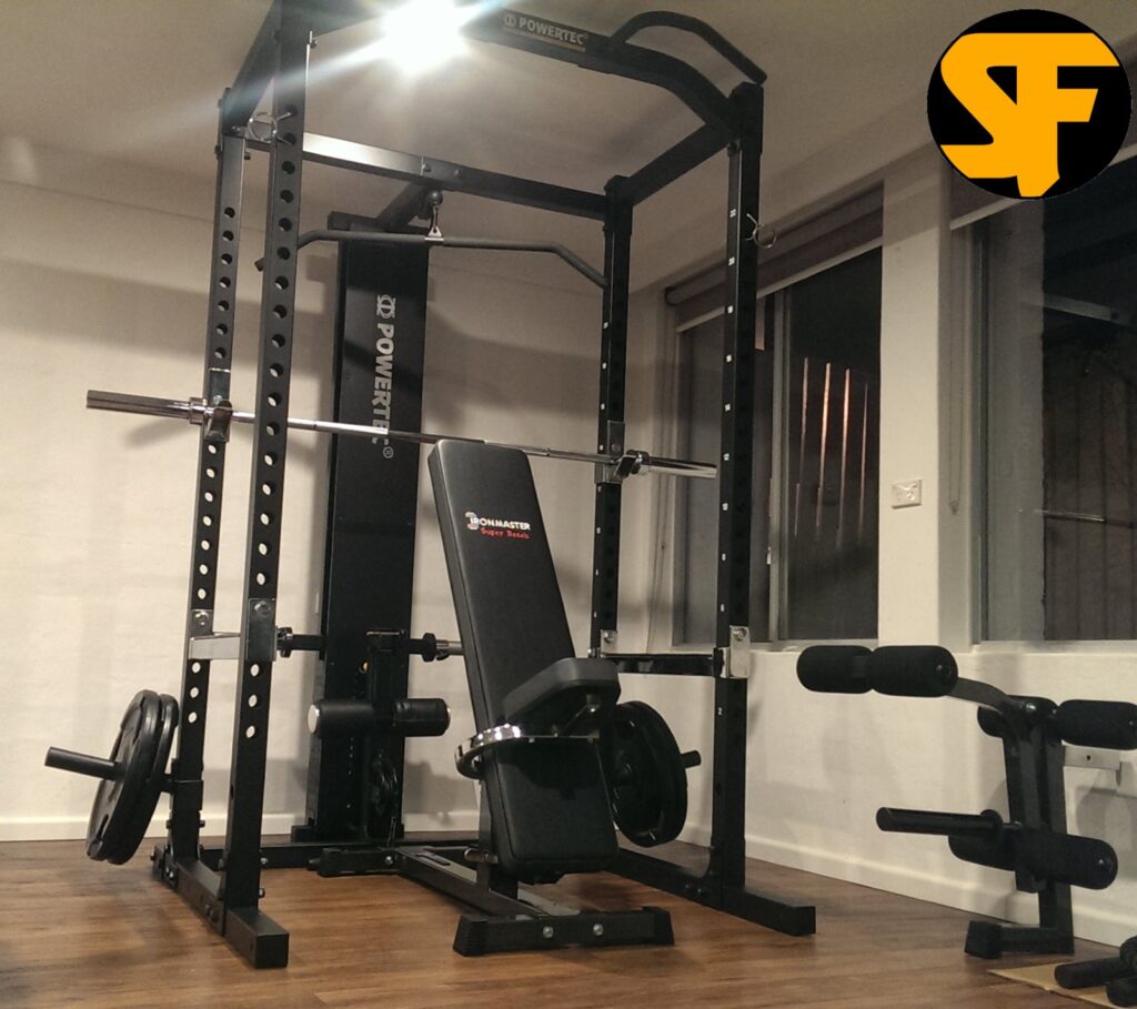 A multi station home gym setup includes a Powertec multigym machine with an adjustable bench press and pull-up bar. The room, adorned with wooden flooring and large windows, features additional exercise equipment like weights and a leg extension pad on the side.