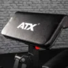 A close-up of a sleek black exercise bench featuring the ATX® logo, set against a backdrop of gray brick. Positioned near the ATX® Bicep Blaster Preacher Curl machine, it is part of a comprehensive gym or fitness equipment setup.