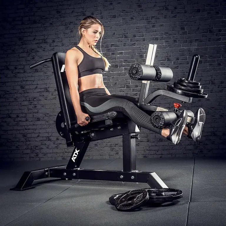 A person is using the ATX Leg Extension Leg Curl Machine in a gym, wearing a black sports bra and leggings. A dark brick wall provides the backdrop, with various weight plates scattered on the floor nearby.