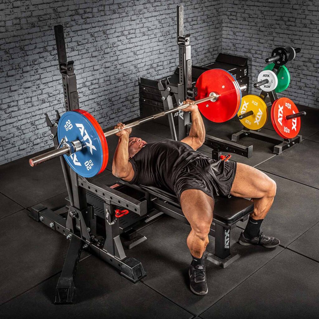 A person uses the ATX® Competition Combo Rack for a bench press, lifting a barbell with weight plates on a sturdy bench. Colorful weight plates are neatly stored on racks against the brick wall behind them.