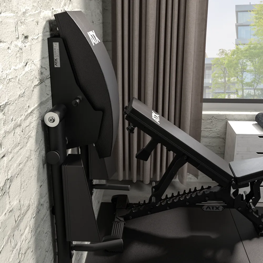 A foldable weight bench is stored upright against a white brick wall in a home gym with gray curtains and a window showing trees and buildings outside, positioned beside the ATX® Wall Mounted Knee Raise & Dip station for versatile workouts.