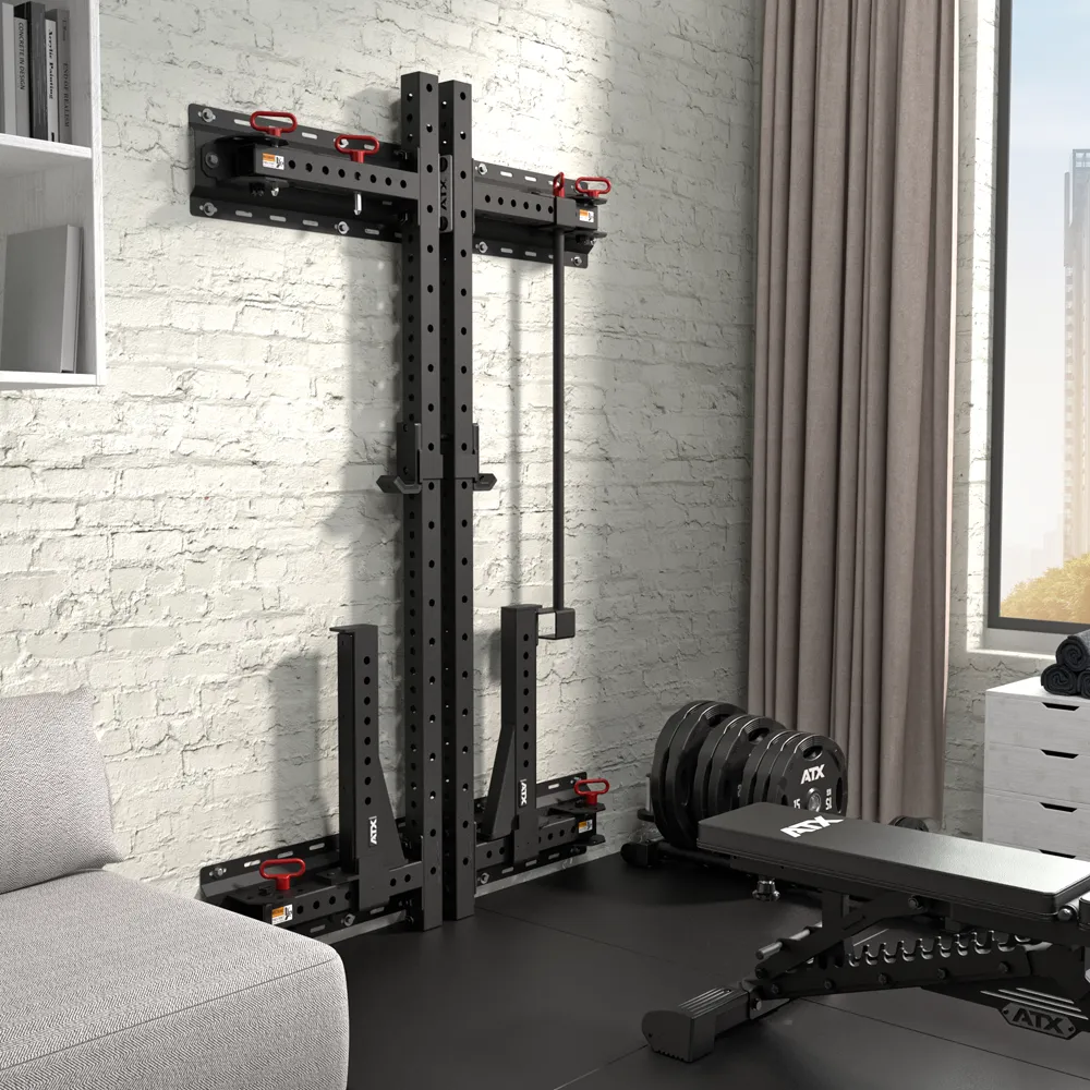 A compact home gym featuring the ATX® Fold Back Rack - Wall Mounted in black, an adjustable weight bench, weight plates, and attachments set against a white brick wall near a window with beige curtains and a gray sofa.