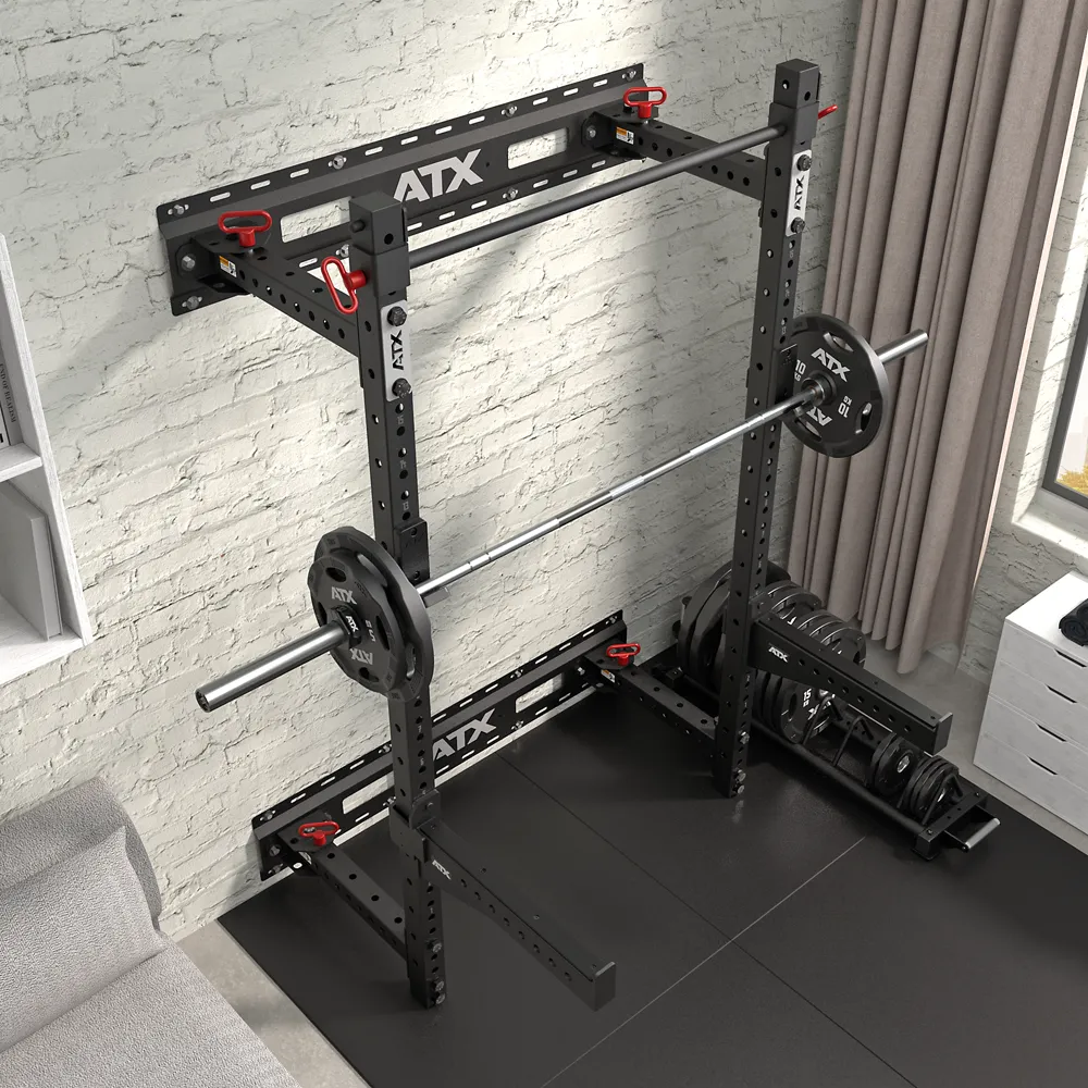An ATX® Fold Back Rack - Wall Mounted with a barbell and weights stands against a white brick wall in a home gym featuring rubber flooring, a dumbbell rack, and a window with curtains.