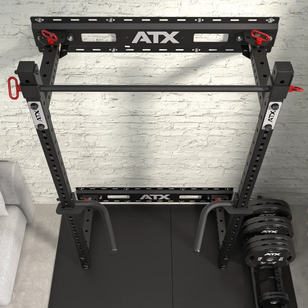 Overhead view of the ATX® Fold Back Rack - Wall Mounted in black, featuring a pull-up bar, weight plates on a storage post, and angled dip bars on a rubber gym mat against a white brick wall. Part of a gray sofa appears on the left.
