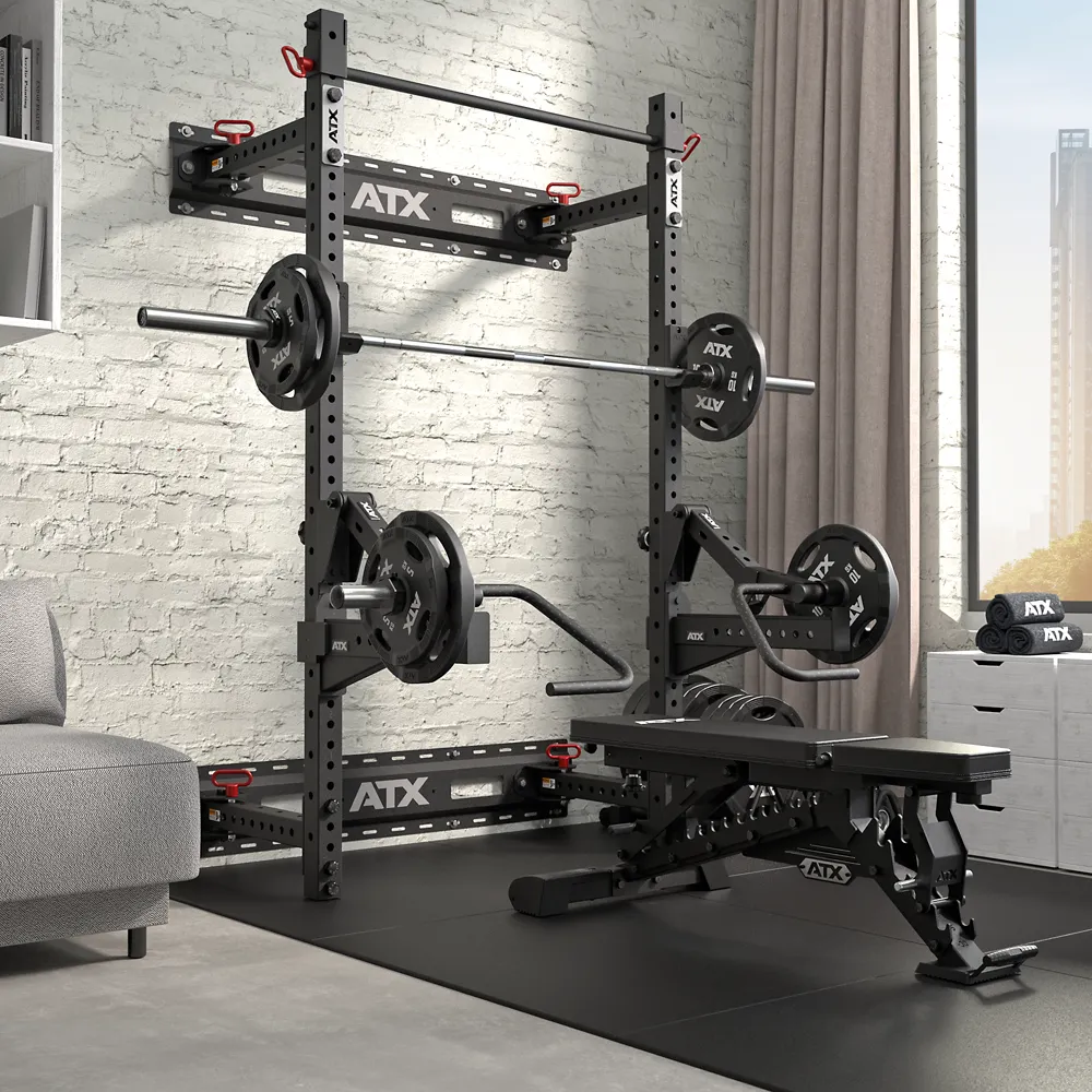 A modern home gym features the ATX® Fold Back Rack - Wall Mounted, loaded barbell, weight plates, adjustable bench, and accessories, all set on black floor mats by a window with city view and gray curtains.