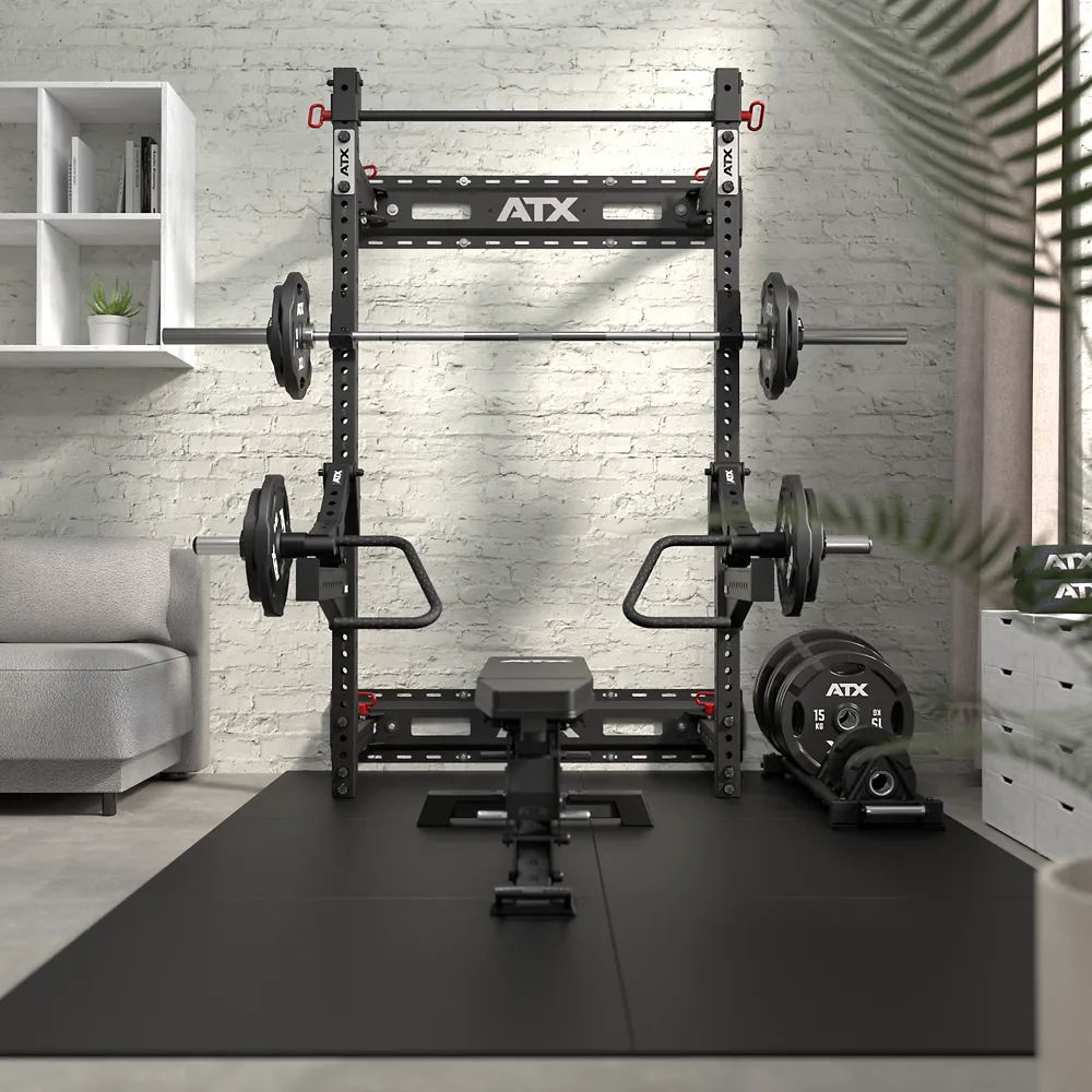 A home gym features the ATX® Fold Back Rack - Wall Mounted with barbells, weight plates, a bench, and gym mats. On the left is a gray sofa and wall shelves; on the right, a large window with curtains lets in natural light.