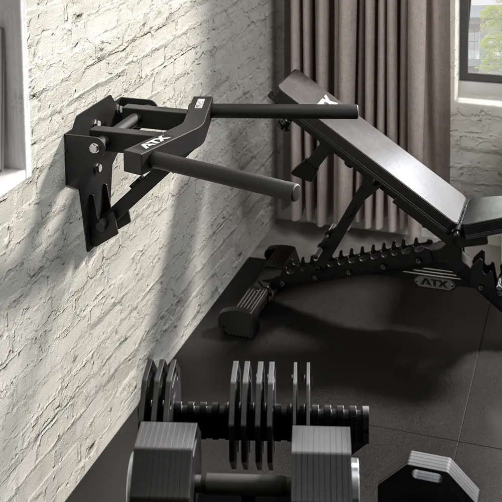 A home gym setup featuring ATX® Folding Wall Mounted Dip Handles, an adjustable weight bench, and a set of dumbbells arranged on the floor against a light brick wall near a window with gray curtains.