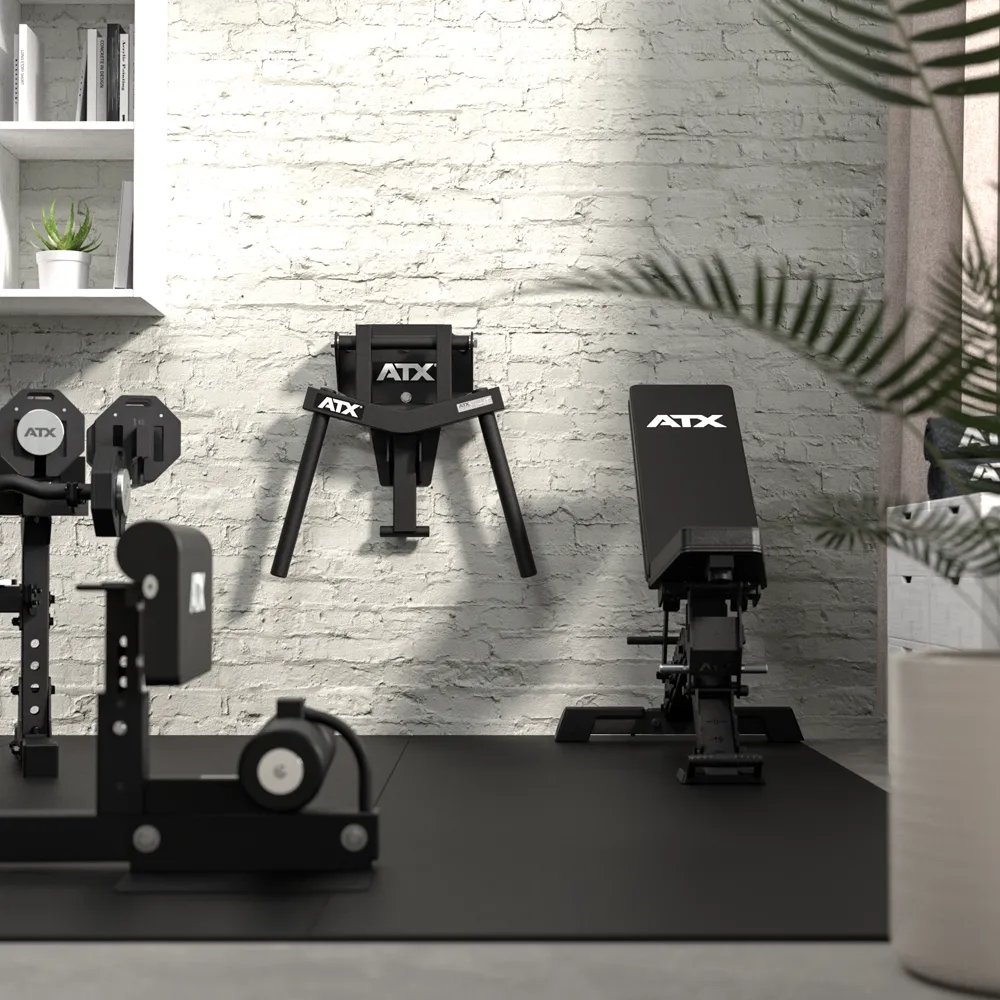 A home gym room featuring ATX workout equipment, including a bench, a wall-mounted machine equipped with ATX® Folding Wall Mounted Dip Handles, and other gear arranged on black mats. White brick walls, a shelf displaying plants, and a leafy plant in the foreground are also visible.