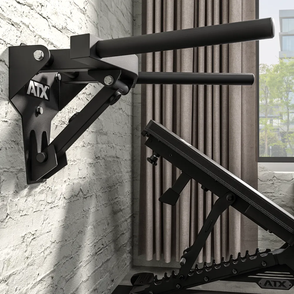 The ATX® Folding Wall Mounted Dip Handles in sleek black are securely mounted to a brick wall in a home gym, with an adjustable weight bench placed below and a window with curtains visible in the background.