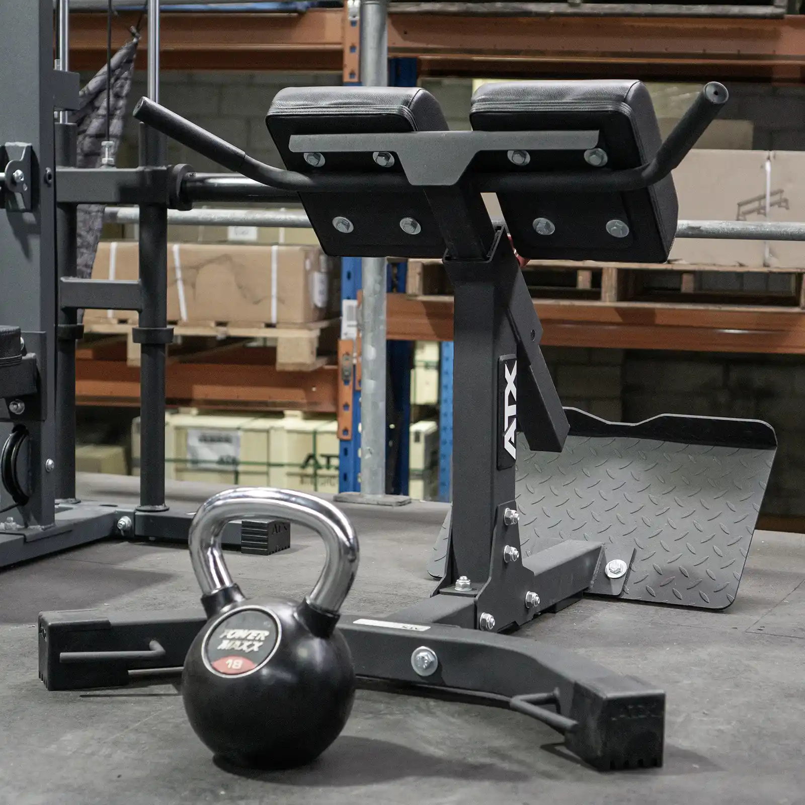 A gym setup featuring the Ironmaster Hyper Core Attachment, commonly referred to as a Roman chair, alongside an 18 kg kettlebell. The equipment rests on a rubber floor, while shelves and cardboard boxes can be seen in the background.